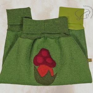 Pode incluir: Cal&ccedil;as de l&atilde; polar verde com uma aplica&ccedil;&atilde;o de cogumelo vermelho e laranja. As cal&ccedil;as t&ecirc;m uma cintura larga e punhos dobrados. O desenho do cogumelo est&aacute; centrado na frente.