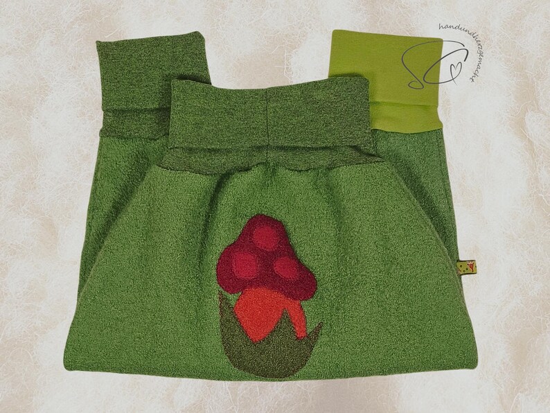 Pode incluir: Cal&ccedil;as de l&atilde; polar verde com uma aplica&ccedil;&atilde;o de cogumelo vermelho e laranja. As cal&ccedil;as t&ecirc;m uma cintura larga e punhos dobrados. O desenho do cogumelo est&aacute; centrado na frente.