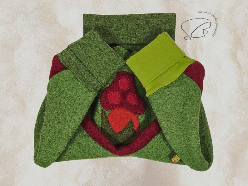 Pode incluir: Um cachecol com capuz verde com uma aplica&ccedil;&atilde;o de cogumelo vermelho. O cachecol &eacute; feito de um tecido macio e texturizado e apresenta um design dobrado com um forro verde claro contrastante. O cogumelo tem um chap&eacute;u e um caule vermelhos.