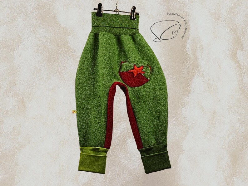 Pode incluir: Cal&ccedil;as de beb&eacute; verdes com um bolso em forma de estrela vermelha e pain&eacute;is interiores das pernas vermelhos. Os punhos e a cintura s&atilde;o de um verde mais escuro. As cal&ccedil;as est&atilde;o penduradas num cabide preto sobre um fundo neutro.
