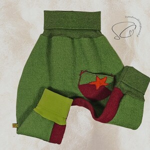 Pode incluir: Cal&ccedil;as de beb&eacute; verdes com um bolso vermelho e verde com uma aplica&ccedil;&atilde;o de estrela. As cal&ccedil;as t&ecirc;m uma cintura alta e punhos. O design inclui uma faixa vermelha na perna. O fundo &eacute; de cor bege claro.