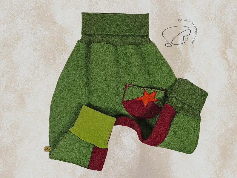 Pode incluir: Cal&ccedil;as de beb&eacute; verdes com um bolso vermelho e verde com uma aplica&ccedil;&atilde;o de estrela. As cal&ccedil;as t&ecirc;m uma cintura alta e punhos. O design inclui uma faixa vermelha na perna. O fundo &eacute; de cor bege claro.