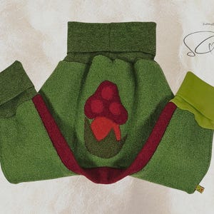 Pode incluir: Um envolt&oacute;rio para beb&eacute; em feltro verde com uma aplica&ccedil;&atilde;o de cogumelo vermelho. O envolt&oacute;rio tem uma faixa vermelha e punhos verdes. Concebido para manter o beb&eacute; quente.