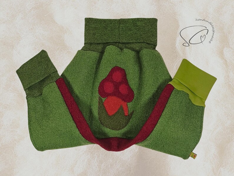 Pode incluir: Um envolt&oacute;rio para beb&eacute; em feltro verde com uma aplica&ccedil;&atilde;o de cogumelo vermelho. O envolt&oacute;rio tem uma faixa vermelha e punhos verdes. Concebido para manter o beb&eacute; quente.