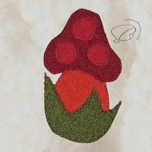 Pode incluir: Aplique de feltro em forma de cogumelo com um chap&eacute;u vermelho com tr&ecirc;s c&iacute;rculos, um caule laranja e folhas verdes. O design feito &agrave; m&atilde;o tem o texto "handmade by SC" em it&aacute;lico.