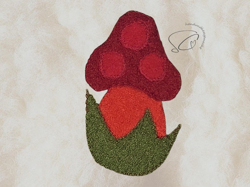 Pode incluir: Aplique de feltro em forma de cogumelo com um chap&eacute;u vermelho com tr&ecirc;s c&iacute;rculos, um caule laranja e folhas verdes. O design feito &agrave; m&atilde;o tem o texto "handmade by SC" em it&aacute;lico.