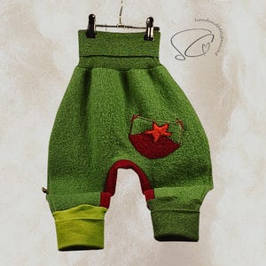 Pode incluir: Cal&ccedil;as de beb&eacute; verdes com um bolso de estrela vermelha e detalhes em vermelho. As cal&ccedil;as s&atilde;o feitas de um tecido texturizado e t&ecirc;m punhos verde lim&atilde;o. As cal&ccedil;as est&atilde;o penduradas num cabide de metal.