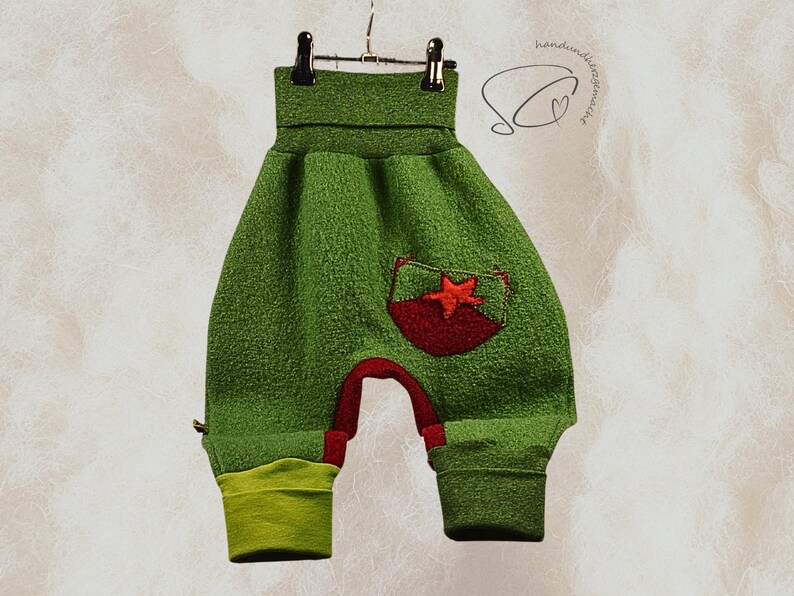 Pode incluir: Cal&ccedil;as de beb&eacute; verdes com um bolso de estrela vermelha e detalhes em vermelho. As cal&ccedil;as s&atilde;o feitas de um tecido texturizado e t&ecirc;m punhos verde lim&atilde;o. As cal&ccedil;as est&atilde;o penduradas num cabide de metal.