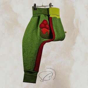 Pode incluir: Cal&ccedil;as de l&atilde; polar verde com uma faixa vermelha na lateral e um bolso vermelho com bagas vermelhas. As cal&ccedil;as t&ecirc;m um punho verde no tornozelo e uma cintura verde lim&atilde;o. As cal&ccedil;as est&atilde;o penduradas num cabide de metal.