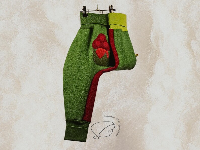 Pode incluir: Cal&ccedil;as de l&atilde; polar verde com uma faixa vermelha na lateral e um bolso vermelho com bagas vermelhas. As cal&ccedil;as t&ecirc;m um punho verde no tornozelo e uma cintura verde lim&atilde;o. As cal&ccedil;as est&atilde;o penduradas num cabide de metal.