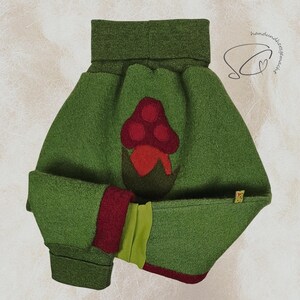 Pode incluir: Um chap&eacute;u de beb&ecirc; de feltro verde com uma aplica&ccedil;&atilde;o de cogumelo vermelho. O chap&eacute;u tem uma aba enrolada e uma guarni&ccedil;&atilde;o vermelha e verde contrastante. O chap&eacute;u foi projetado para manter a cabe&ccedil;a do beb&ecirc; aquecida.
