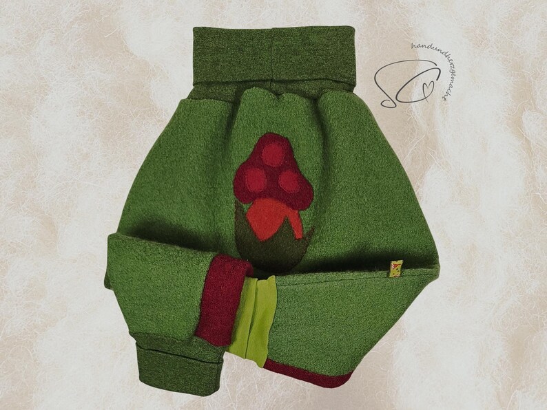 Pode incluir: Um chap&eacute;u de beb&ecirc; de feltro verde com uma aplica&ccedil;&atilde;o de cogumelo vermelho. O chap&eacute;u tem uma aba enrolada e uma guarni&ccedil;&atilde;o vermelha e verde contrastante. O chap&eacute;u foi projetado para manter a cabe&ccedil;a do beb&ecirc; aquecida.