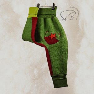 Pode incluir: Cal&ccedil;as de l&atilde; polar verde com uma faixa vermelha na lateral e um bolso de estrela vermelha. As cal&ccedil;as t&ecirc;m punho e c&oacute;s verdes com um detalhe verde lim&atilde;o. As cal&ccedil;as est&atilde;o penduradas num cabide.