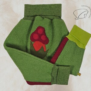 Pode incluir: Roupa de l&atilde; feltrada verde com uma aplica&ccedil;&atilde;o de cogumelo. A roupa apresenta uma gola alta e punhos em um verde mais claro. Um cogumelo vermelho com um caule vermelho escuro est&aacute; centrado na frente. As mangas t&ecirc;m uma faixa vermelha na lateral.