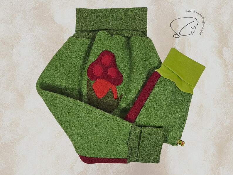 Pode incluir: Roupa de l&atilde; feltrada verde com uma aplica&ccedil;&atilde;o de cogumelo. A roupa apresenta uma gola alta e punhos em um verde mais claro. Um cogumelo vermelho com um caule vermelho escuro est&aacute; centrado na frente. As mangas t&ecirc;m uma faixa vermelha na lateral.