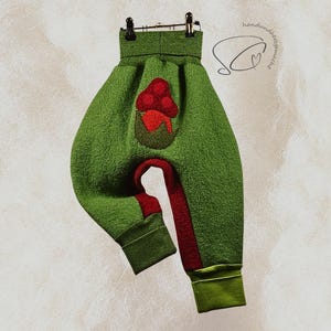 Pode incluir: Cal&ccedil;as de beb&eacute; verdes com uma aplica&ccedil;&atilde;o de cogumelo vermelho. As cal&ccedil;as t&ecirc;m um tecido texturizado, detalhes vermelhos nas pernas e punhos verdes. As cal&ccedil;as est&atilde;o penduradas num cabide preto.