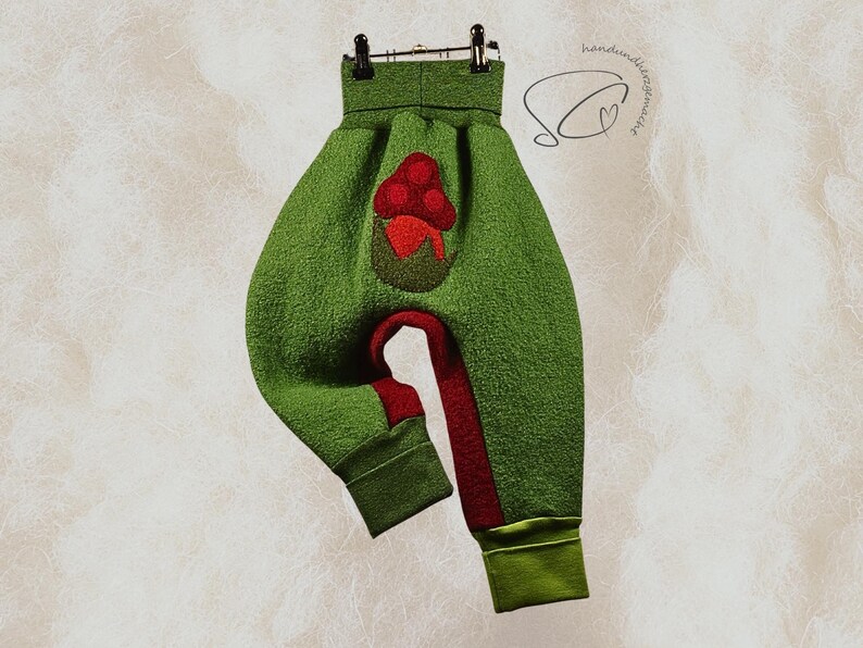 Pode incluir: Cal&ccedil;as de beb&eacute; verdes com uma aplica&ccedil;&atilde;o de cogumelo vermelho. As cal&ccedil;as t&ecirc;m um tecido texturizado, detalhes vermelhos nas pernas e punhos verdes. As cal&ccedil;as est&atilde;o penduradas num cabide preto.