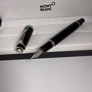 Op de afbeelding: Een zwarte Montblanc vulpen met een zilverkleurige penpunt en een bijpassende dop met een blauwe edelsteen op de clip. De pen wordt gepresenteerd in een doos met witte voering en het Montblanc-logo gedrukt op de binnenkant van het deksel.