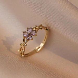 May include: A gold-colored ring featuring a four-petal flower design with purple gemstone petals. The ring has a delicate, adjustable band and is set against a soft, neutral background.