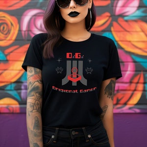 Puede incluir: Una camiseta negra con un diseño gráfico que presenta un joystick de videojuego retro y el texto "O.G. Original Gamer".