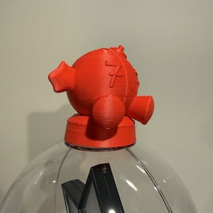 May include: A bright orange 3D-printed spherical topper with a textured surface, featuring a prominent number 7 embossed on its side. The object has several protruding rounded elements and is positioned atop a clear, spherical container.