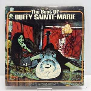 May include: Album cover for "The Best Of Buffy Sainte-Marie." The cover features a woman with long dark hair, reclining with a silver guitar. The background includes red curtains and a patterned rug. The album title is in white text.