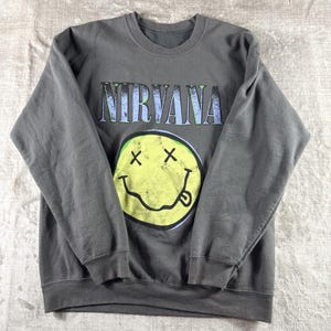 May include: Gray sweatshirt featuring the band name "NIRVANA" in a distressed font above a yellow smiley face with crossed-out eyes and a protruding tongue. The sweatshirt is a crew neck style.