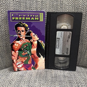 May include: A VHS tape and its cover for the movie "Crying Freeman." The cover is purple with the movie title in yellow and green. The cover art features a man with a dragon tattoo and a woman. The VHS tape is black.