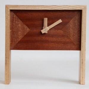 May include: A wooden desk clock with a rectangular frame and a geometric design. The clock face is a dark brown colour, with a lighter wood frame and clock hands. The clock stands on two rectangular legs.