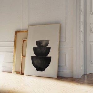 May include: A black and white minimalist art print featuring three stacked bowls. The print is framed in a thin gold frame and is leaning against a white wall.