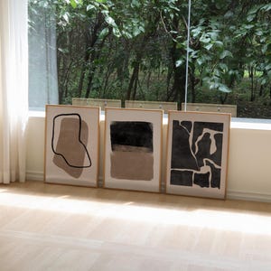 May include: Three framed abstract art prints with black and brown shapes on a white background. The prints are displayed on a light wood floor in front of a window with a view of trees.