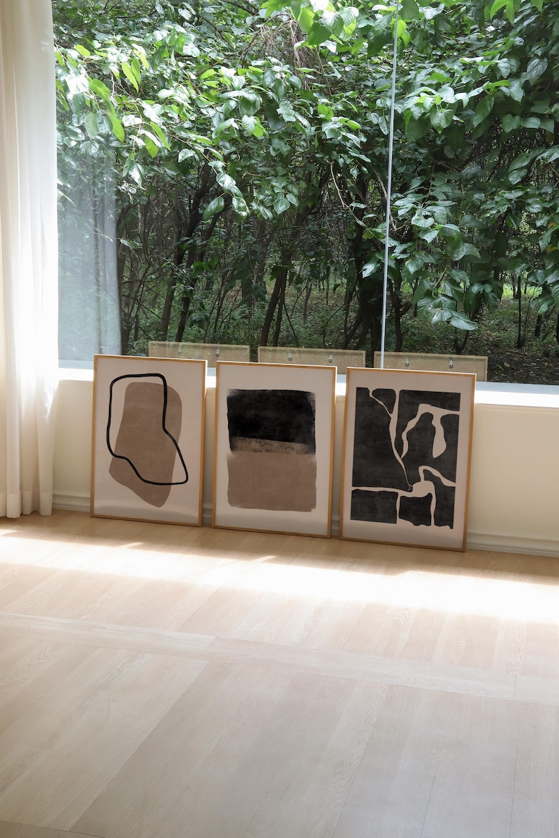 May include: Three framed abstract art prints with black and brown shapes on a white background. The prints are displayed on a light wood floor in front of a window with a view of trees.