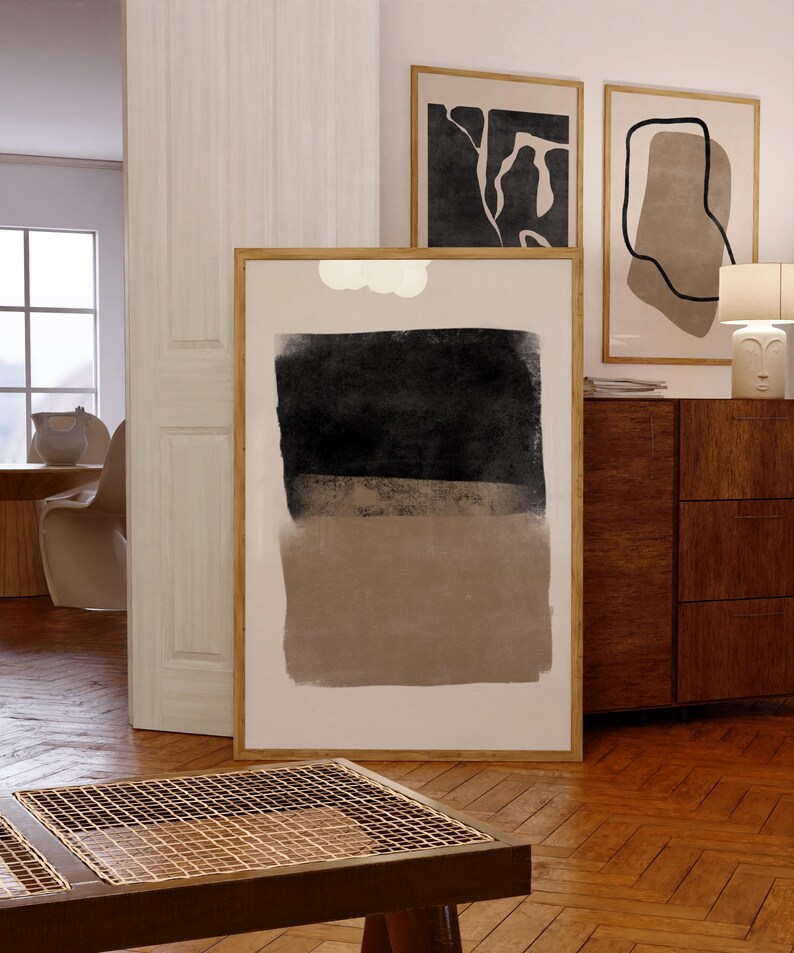 May include: Three framed abstract art prints with black and brown shapes on a white background. The prints are hanging on a white wall above a wooden dresser.