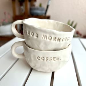 Puede incluir: Dos tazas de café de cerámica blanquecina apiladas. La taza superior dice "GOOD MORNING" y la inferior "COFFEE". Ambas tazas tienen una superficie texturizada y un asa.