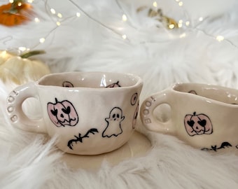 May include: Two white ceramic mugs with black and pink Halloween themed designs. The mugs have the word "BOO!" on the handles. One mug has a ghost, a bat, and a pumpkin with a pink heart. The other mug has a pumpkin with a pink heart and a bat.