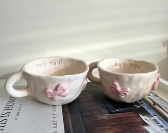 May include: Two off-white ceramic teacups with a pink bow accent and the text "FOR YOU". The cups have a handle and are placed on a book with the text "THIS IS HOME".