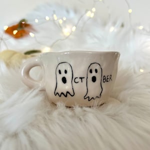 Könnte beinhalten: Weiße Keramiktasse mit zwei Geisterillustrationen und dem Text "OCT BER" auf der Vorderseite.