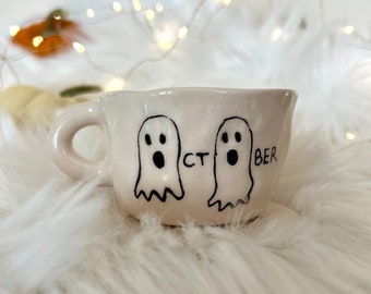 May include: White ceramic mug with two ghost illustrations and the text "OCT BER" on the front.