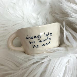 May include: A small, off-white ceramic mug with a curved handle. The mug features the handwritten phrase "always late but worth the wait" in dark blue ink. The mug is set against a fluffy, white background.
