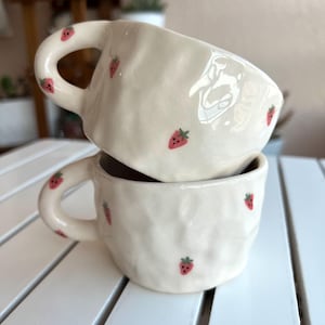 Könnte beinhalten: Zwei gestapelte, cremefarbene Keramikbecher mit strukturierter Oberfläche. Jeder Becher ist mit kleinen, handgemalten roten Erdbeeren mit grünen Blättern verziert. Die Becher haben abgerundete Henkel und stehen auf einer weißen Holzoberfläche.