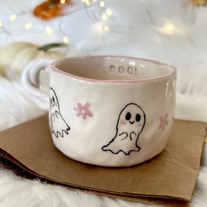 May include: A white ceramic mug with a pink rim and the word "BOO!" written on it. The mug is decorated with two ghosts and pink flowers.