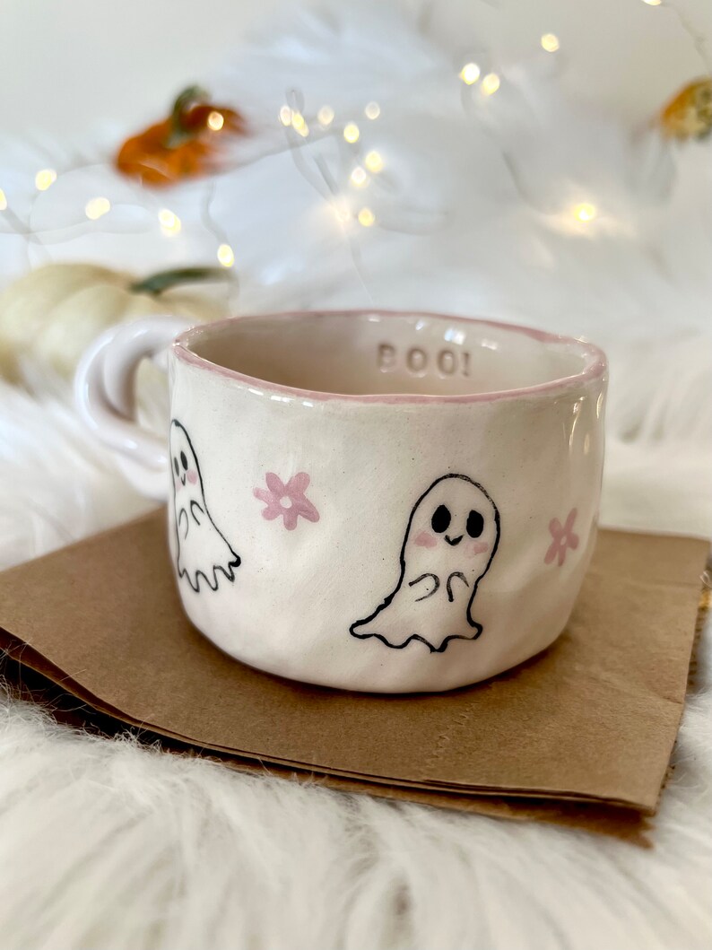 May include: A white ceramic mug with a pink rim and the word "BOO!" written on it. The mug is decorated with two ghosts and pink flowers.
