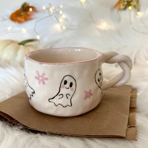 May include: A white ceramic mug with a pink rim and a whimsical design of ghosts and pink flowers. The mug has a twisted handle.