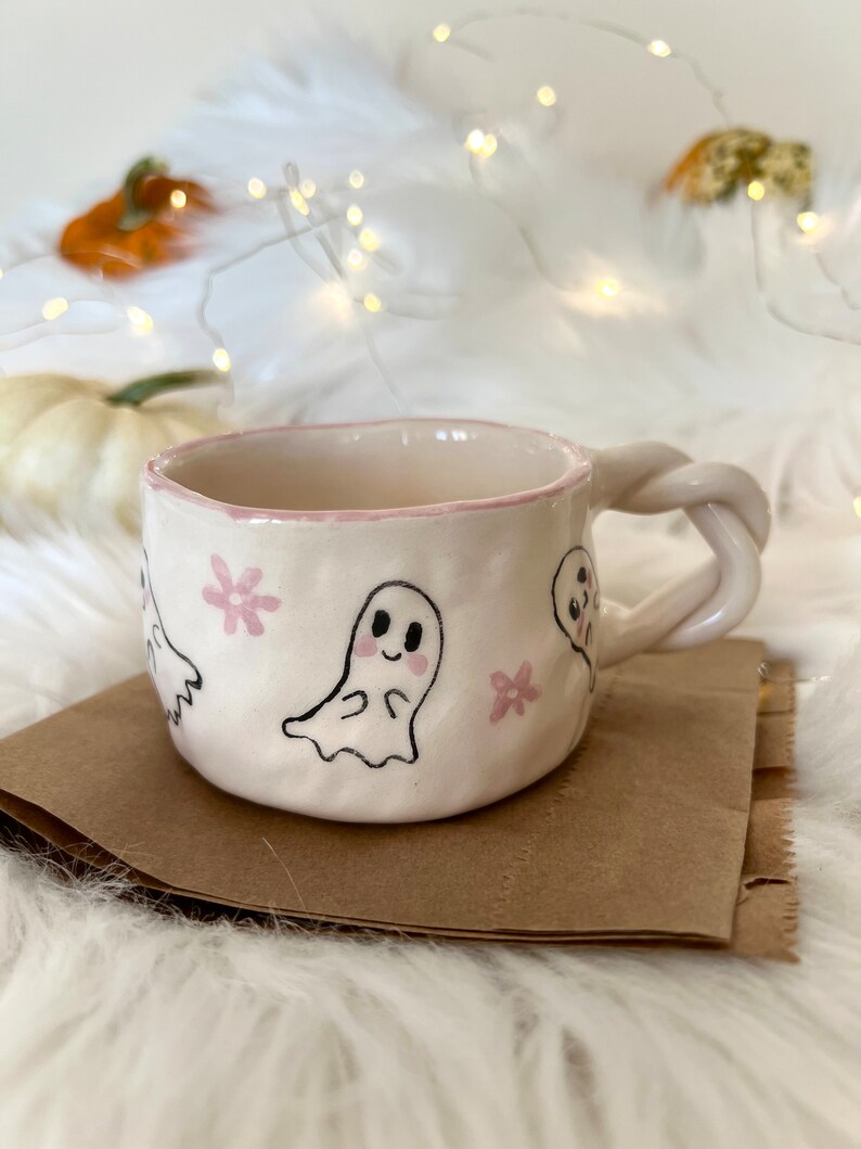 May include: A white ceramic mug with a pink rim and a whimsical design of ghosts and pink flowers. The mug has a twisted handle.
