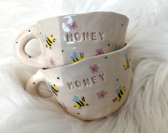 May include: Two white ceramic honey pots with a bee and flower design. The pots have the word "Honey" written on them.