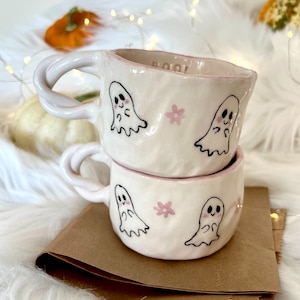 Può includere: Due tazze in ceramica bianca con accenti rosa e illustrazioni di fantasmi. Le tazze sono impilate l'una sull'altra. La tazza superiore ha la parola "Boo!" scritta sul lato.