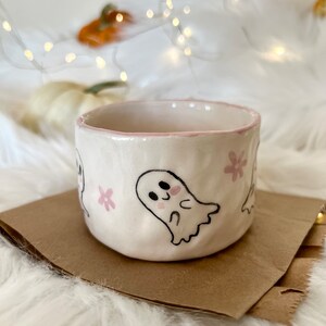 May include: A white ceramic mug with a pink rim and pink flower designs. The mug features a hand-painted design of a cute ghost with blush cheeks.