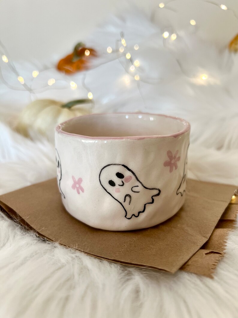May include: A white ceramic mug with a pink rim and pink flower designs. The mug features a hand-painted design of a cute ghost with blush cheeks.