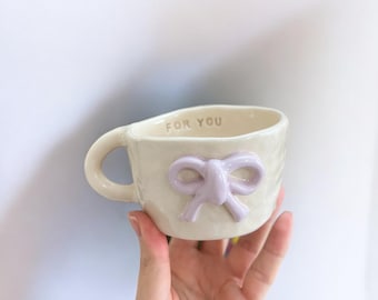 May include: A white ceramic mug with a raised purple bow and the words "For You" on the side.