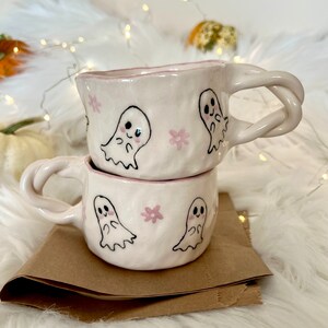 May include: Two white ceramic mugs with pink floral and ghost designs. The mugs are stacked on top of each other. The mug on top has a twisted handle.
